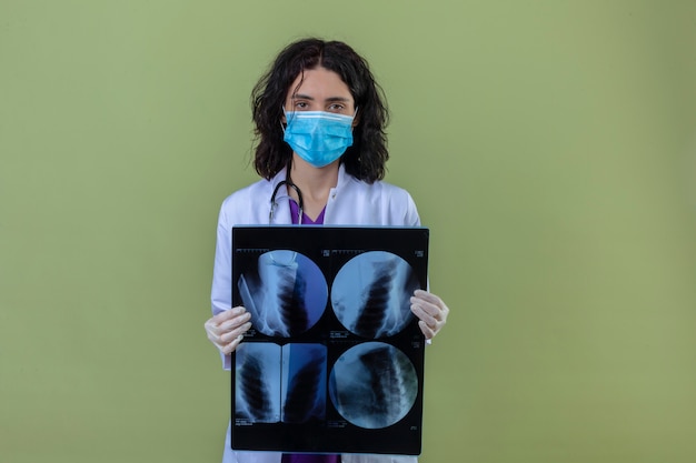 Free photo woman doctor wearing white coat with stethoscope in medical protective mask standing with x-ray of lungs with serious face on isolated green