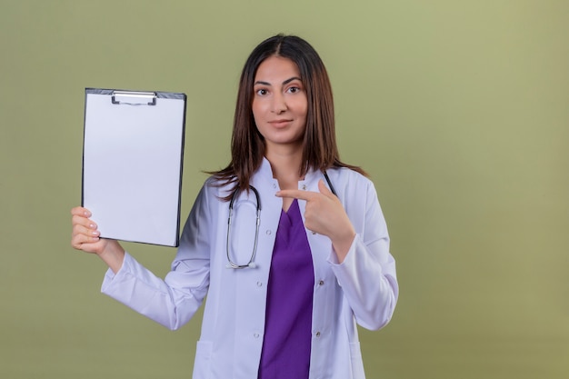 woman doctor wearing white coat and with stethoscope pointing with index finger to clipboard in hand standing on isolated green