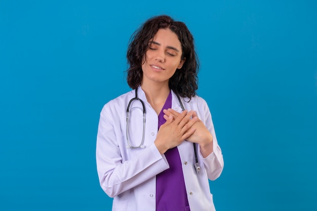 woman doctor wearing white coat with stethoscope smiling with hands on chest with closed eyes and grateful gesture on face standing on isolated blue