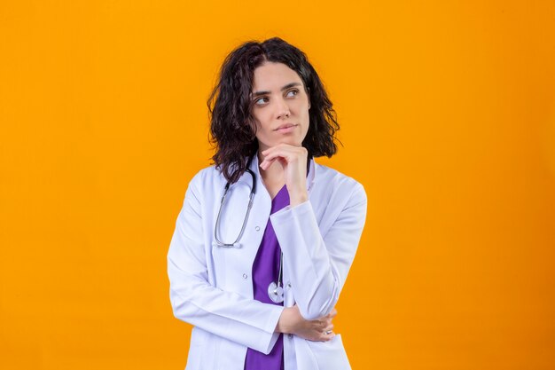 женщина-врач в белом халате со стетоскопом, стоя с рукой на подбородке, глядя в сторону, думая, мечтательный взгляд, стоя на изолированном апельсине