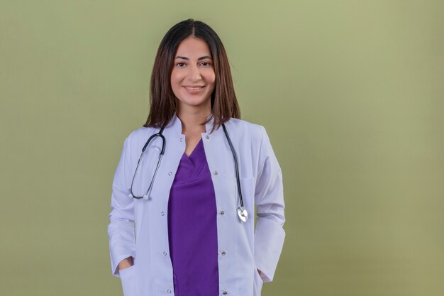 woman doctor wearing white coat and with stethoscope standing with hands in pockets smiling confident looking positive and happy on isolated green