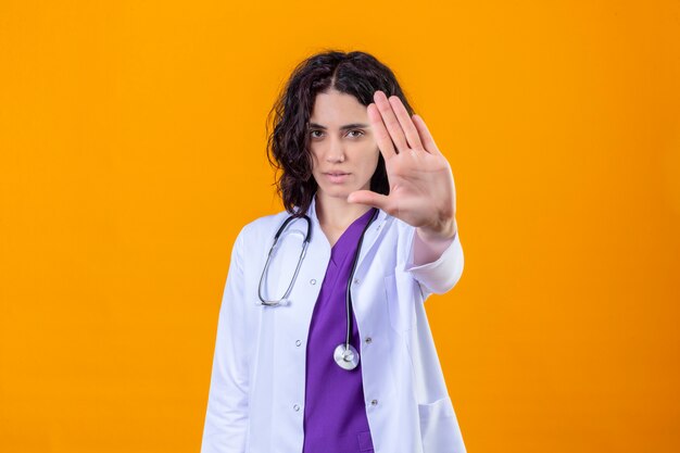 woman doctor wearing white coat with stethoscope standing with open hand doing stop sign with serious and confident expression defense gesture on isolated orange