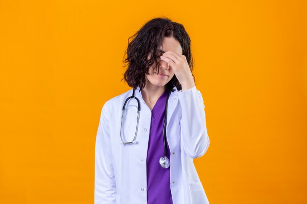 woman doctor wearing white coat with stethoscope touching nose between closed eyes stressed feeling fatigue standing on isolated orange