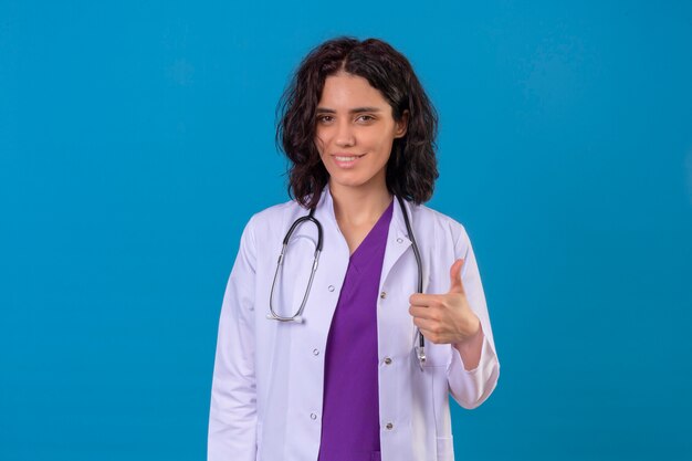 격리 된 파란색에 서 엄지 손가락을 보여주는 행복 한 미소와 청진 기 흰색 코트를 입고 여자 의사