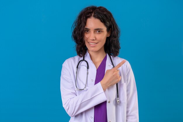 woman doctor wearing white coat with stethoscope with smile on face pointing with finger to the side standing on isolated blue