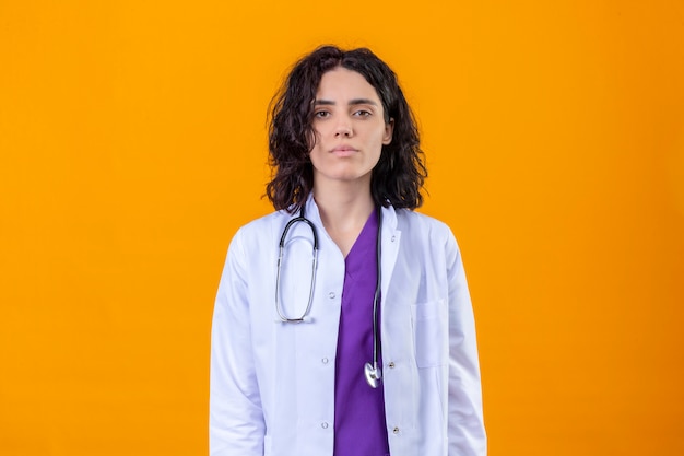 woman doctor wearing white coat with stethoscope without smile looking confident standing on isolated orange