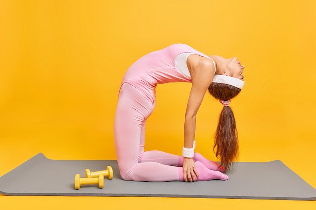 Free photo woman does pilates on fitness mat has perfect slim figure leans backwards wears headband and activewear exercises with dumbbells isolated on yellow