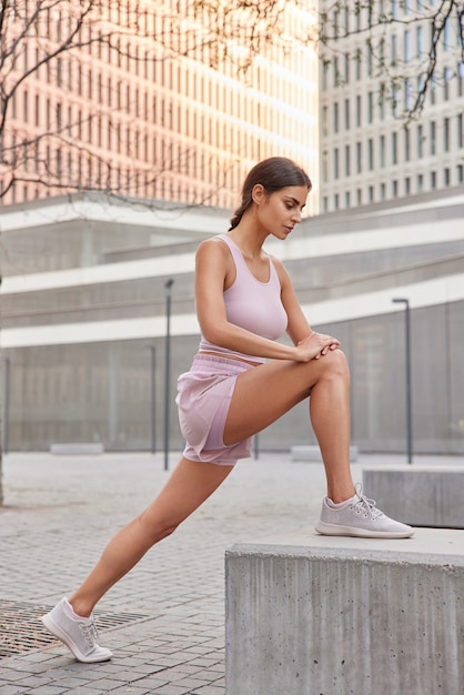  woman does sport exercises outdoors stretches legs warms up before jogging wears cropped top shorts and sneakers poses on city establishments