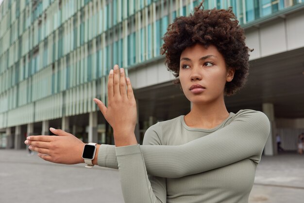 La donna fa esercizi di riscaldamento allunga le braccia prima dell'allenamento indossa smartwatch e tuta sportiva guarda seriamente in pose a distanza contro il moderno edificio della città