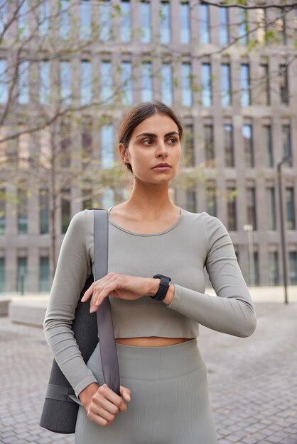 woman dressed in activewear checks time on smartwatch carries fitness mat poses against modern city building going to have training looks thoughtfully somewhere