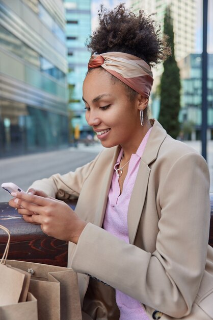 woman dressed in beige formal jacket headband uses mobile phone chats online uses wifi connection poses at urban setting with paper bags around