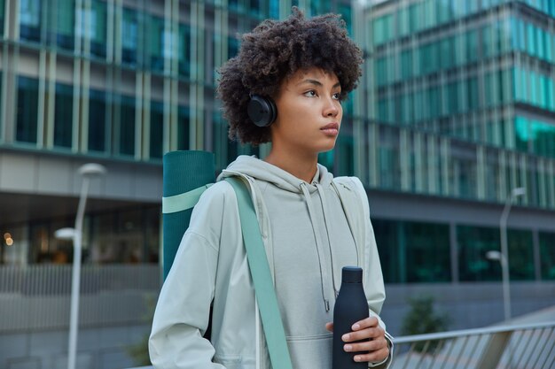 woman dressed in casual hoodie carried karemat for fitness training drinks fresh water listens music has regular exercising in urban setting