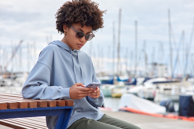 無料写真 カジュアルなパーカーの流行のサングラスに身を包んだ女性はスマートフォンの小切手を使用します通知は港の近くの木製のベンチに座って船やヨットの景色を賞賛します