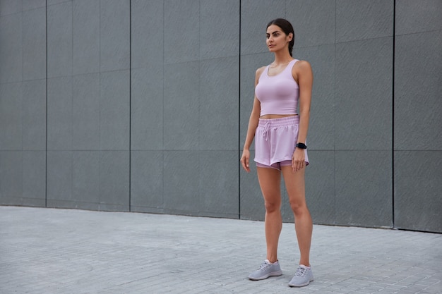 woman dressed in sportswear wears smartwatch focused into distance has cardio workout for keeping fit stands against grey wall