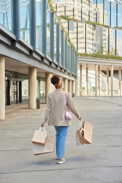 woman dressed in stylish jacket jeans and sportshoes carries many paper bags returns home after making successful shopping poses in downtown around modern glass buildings