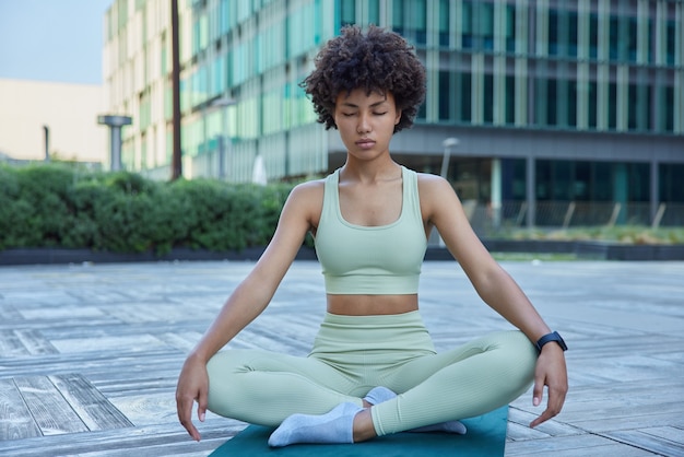 Woman dressed in track suit meditates on fitness mat sits crossed legs poses in downtown against modern building tries to relieve negative emotions