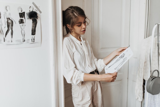 Woman dressed in white outfit looks at clothing samples Darkhaired lady in light blouse and pants poses in cozy fashion designer office