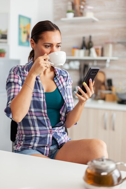 Woman drinking aromatic tea in the morning browsing on smartphone