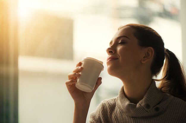 窓を通ってストリーミングしてレンズにフレアを作り出す、日の出と一緒にコーヒーを飲む女性。