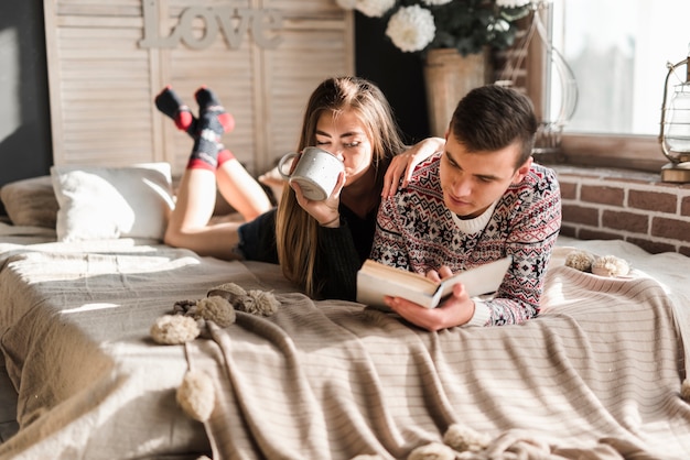 Woman drinking coffee lying with her boyfriend reading book on bed