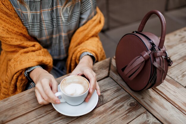 Woman drinking coffee . Stylish bag on table. Wearing grey dress and orange plaid. Enjoying cozy morning in cafe.