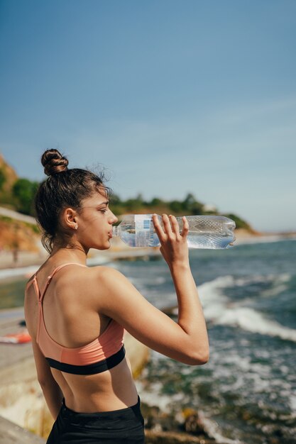 женщина пьет пресную воду из бутылки после тренировки на пляже