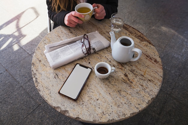Женщина пьет чай за столом с газетой и смартфоном