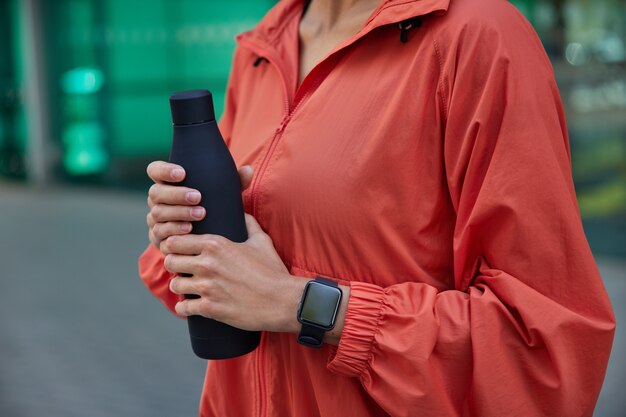  woman drinks water after hard training outdoors holds bottle of water uses smartwatch dressed in windbreaker feels thirsty after sport practice poses on blurred 