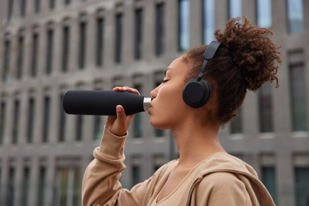 woman drinks water during workout break dressed inn sportsclothes listens audio track in wireless headphones poses outside