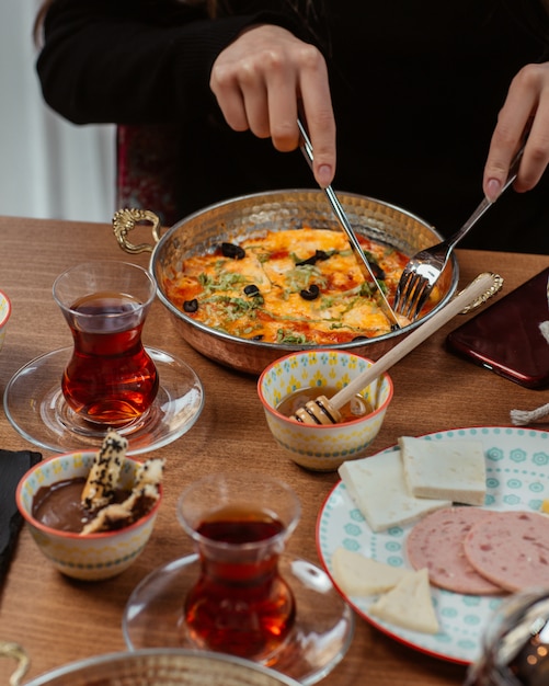Женщина ест завтрак омлет внутри сковороды, вокруг стола, подаренного медом, сыром и салями и черным чаем.