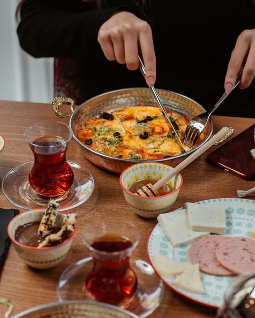 蜂蜜、チーズ、サラミ、紅茶を寄付したテーブルの周りで、パンの中で朝食オムレツを食べる女性。