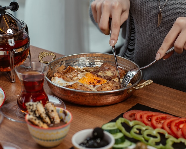 A woman eating breakfast omlette inside a pan, around a table donated with olives, vegetables and black tea.