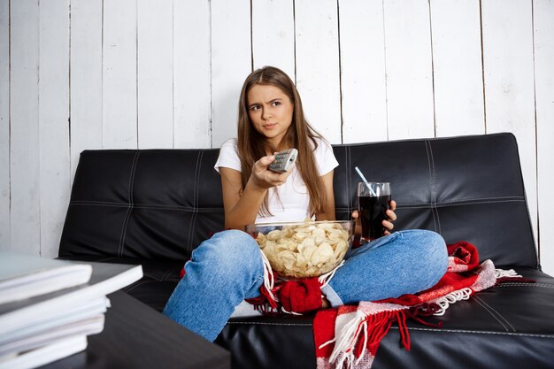 woman eating chips, drinking soda, watching tv, sitting on sofa.
