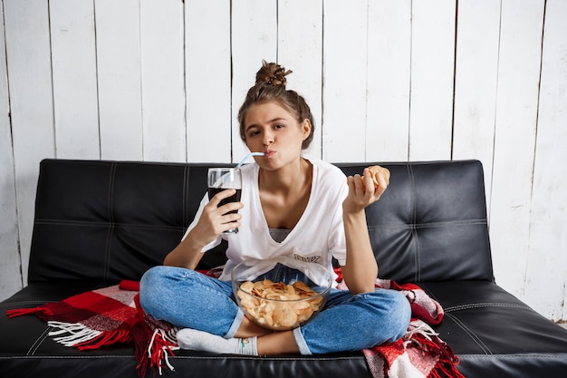 woman eating chips, drinking soda, watching tv, sitting at sofa.