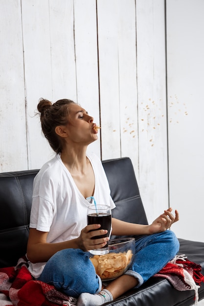 チップを食べる、ソーダを飲む、テレビを見る、ソファーに座っている女性。