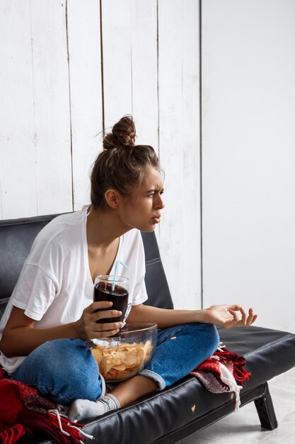 woman eating chips, drinking soda, watching tv, sitting at sofa.