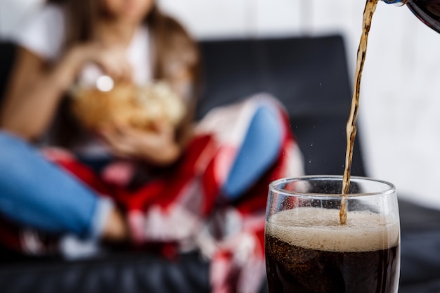 woman eating chips, sitting on sofa. Fosuc  glass of soda.