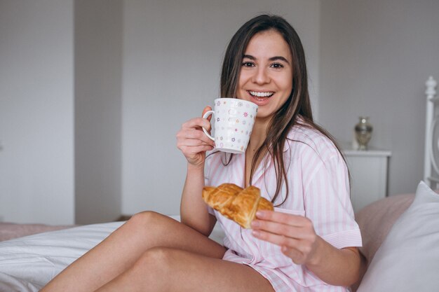 Woman eating delicious croissant with coffee in bed