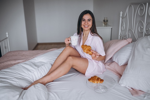 Woman eating delicious croissant with coffee in bed