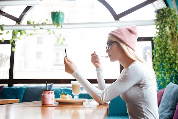 Женщина ест десерт и использует мобильный телефон в кафе