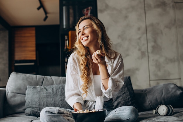 Женщина ест попкорн дома на диване и смотрит фильм