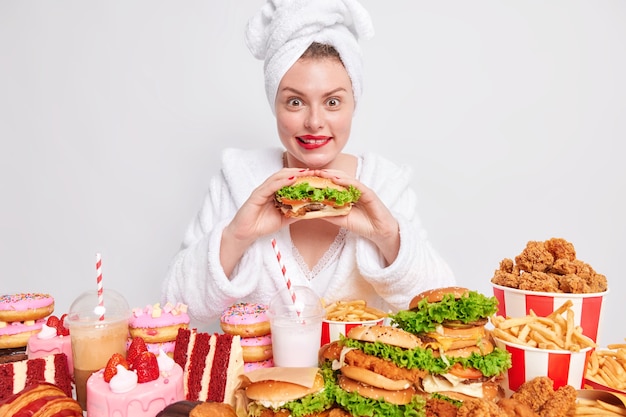 woman eats greedily burger likes cheat meal and unhealthy junk food has oneating habit wears bathrobe and towel on head surrounded by various tasty treaats on white