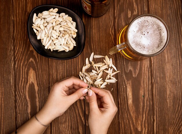 A woman eats salty snack sunflower seeds with a mug of beer on rustic wood top view