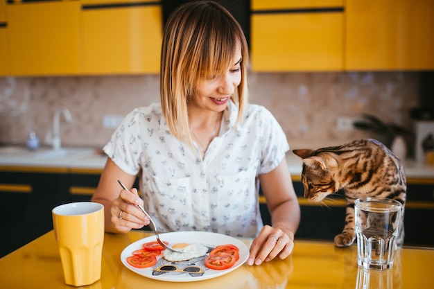 Woman eats at the table while Bengla cat stands behind her 