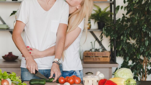 キッチンカウンターでナイフで野菜を切る彼女のボーイフレンドを抱きしめている女性