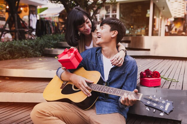 Woman embracing a man and holding a gift while he plays the guitar