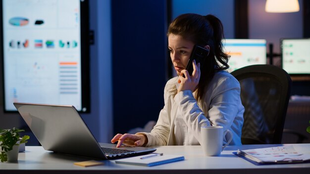 夜遅くにラップトップで働いている間電話で話す女性従業員。忙しい集中フリーランサーは、仕事の読み書き、検索、休憩のために残業をしている最新のテクノロジーネットワークワイヤレスを使用しています