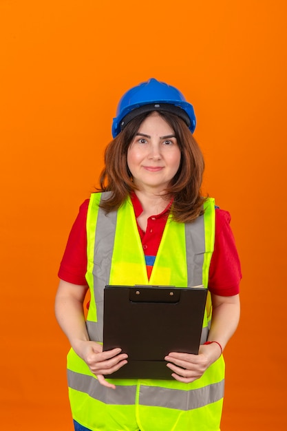 Ingegnere della donna in maglia della costruzione e lavagna per appunti della tenuta del casco di sicurezza in mani che sembrano sicure con il sorriso sul fronte che controlla parete arancio isolata