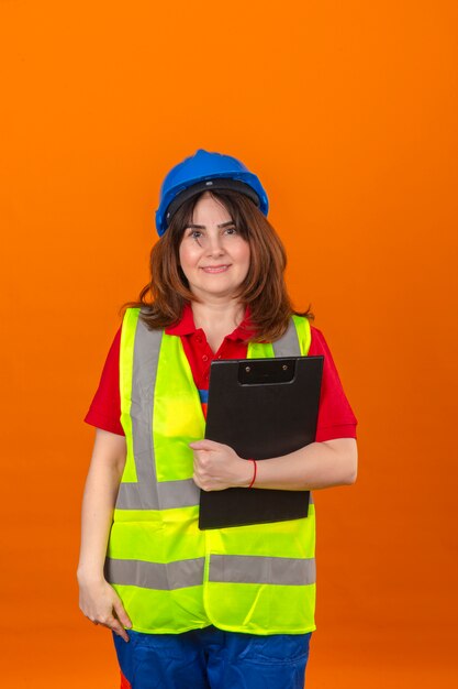Женщина-инженер в строительном жилете и защитном шлеме, держа в руках буфер обмена, выглядит уверенно с улыбкой на лице, стоя над изолированной оранжевой стеной