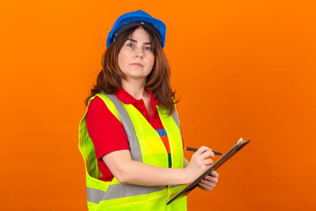 Женщина-инженер в строительном жилете и защитном шлеме держит буфер обмена с ручкой в руках, выглядит уверенно и гордо, стоя над изолированной оранжевой стеной